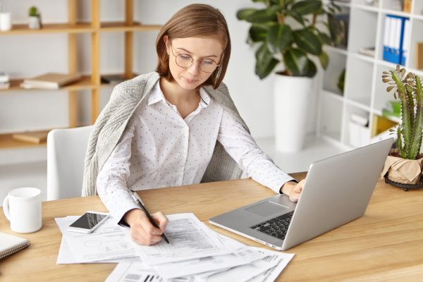 Serious woman wears formal white blouse, works in office, fills in business documents, surrounded with laptop and smart phone. Female boss prepares necessary papers for presenting on meeting Serious woman wears formal white blouse, works in office, fills in business documents, surrounded with laptop and smart phone. Female boss prepares necessary papers for presenting on meeting